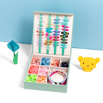 The hair accessory organiser in mint green is filled with hair accessories and is placed open on a white surface with a blue backdrop. It is accessorised with some origami craft.