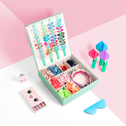 Mint green hair accessory storage box is shown against a pink and white background. The image shows the box with hair clips attached to the internal ribbons, and hair bobbles, head bands and scrunchies are neatly organised in the bottom grid compartment. There are origami paper works surrounding it and a cute girls pen set.