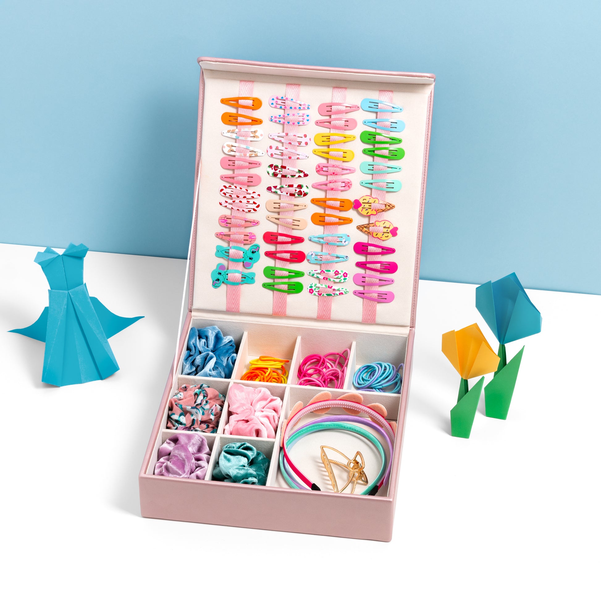 The hair accessory organiser in pink is filled with hair accessories and is placed open on a white surface with a blue backdrop. It is accessorised with some origami art work.