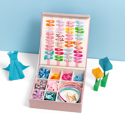 The hair accessory organiser in pink is filled with hair accessories and is placed open on a white surface with a blue backdrop. It is accessorised with some origami art work.