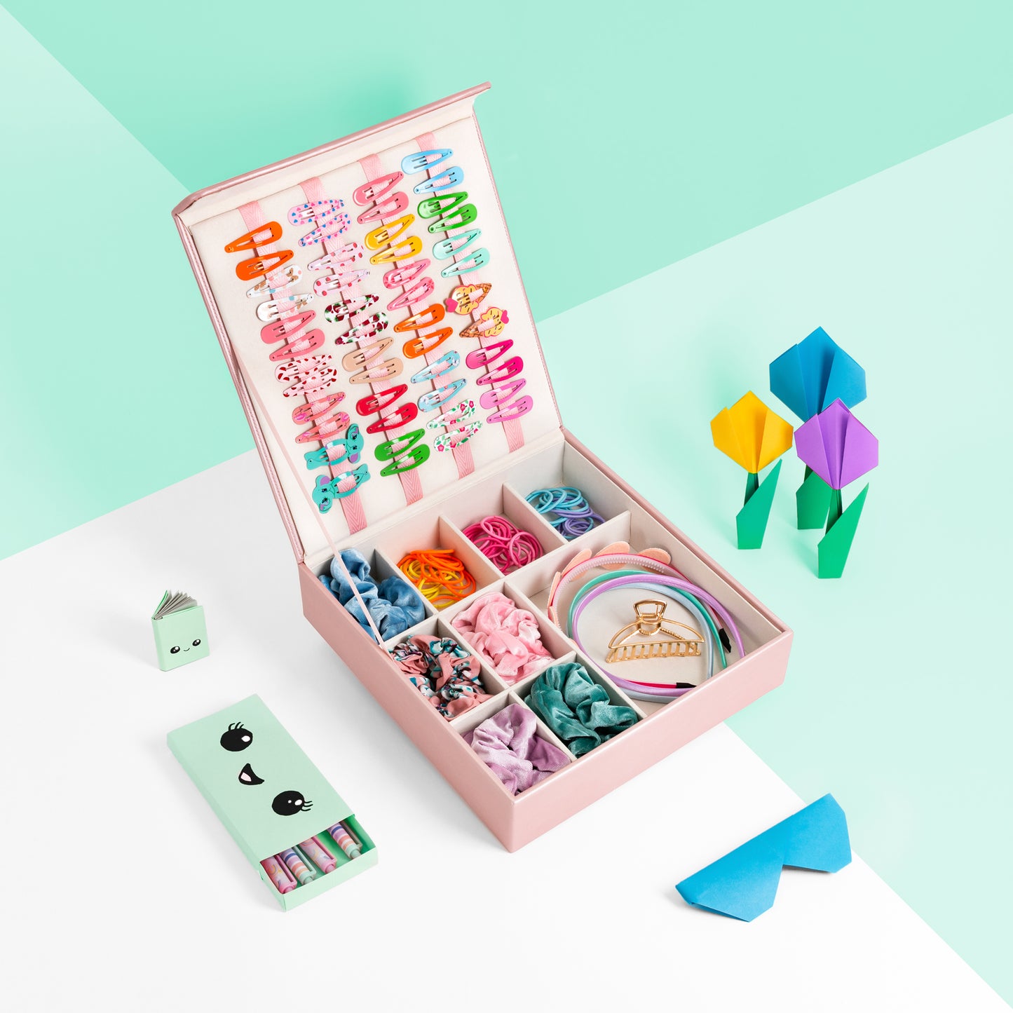 Pink hair accessory storage box is shown against a mint green and white background. The image shows the box with hair clips attached to the internal ribbons, and hair bobbles, head bands and scrunchies are neatly organised in the bottom grid compartment. There are origami paper works surrounding it and a cute girls pen set